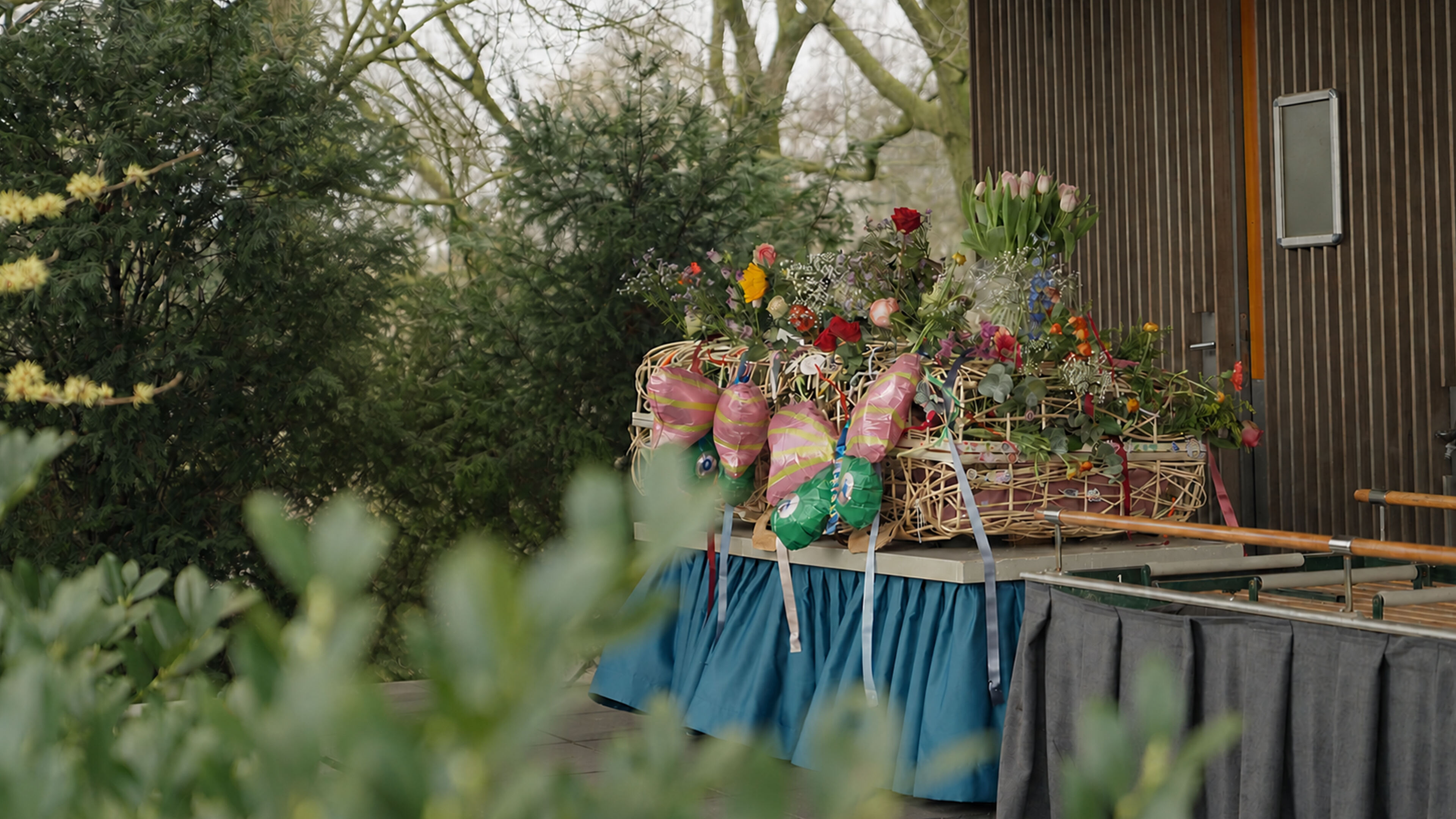 Bloemen en ballonnen op een uitvaartkist tijdens een afscheid in de buitenlucht.