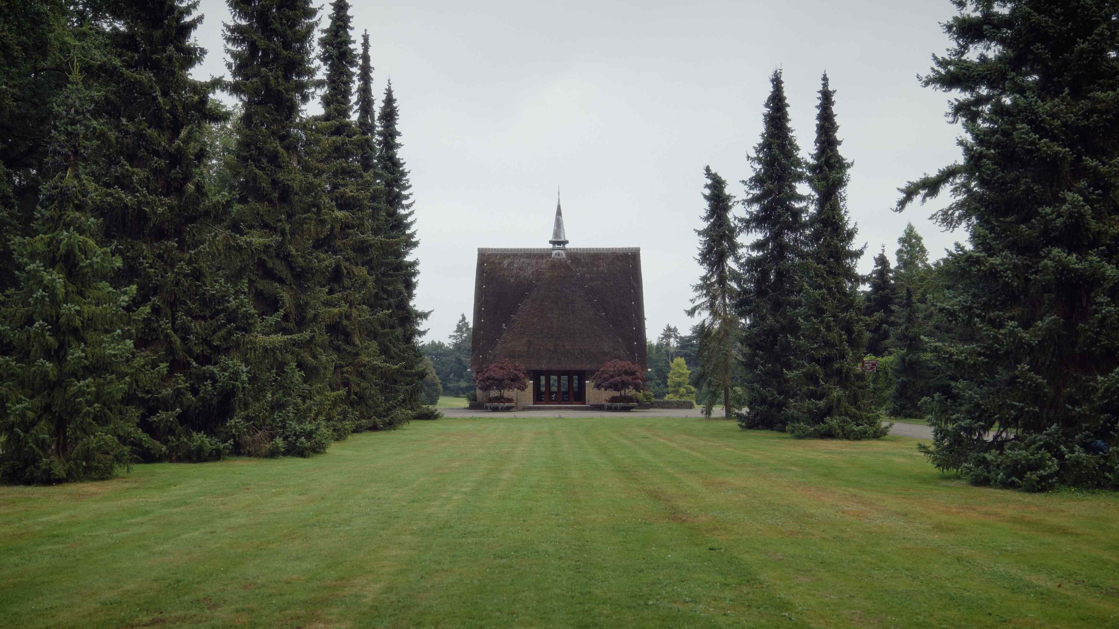 Kapel op een begraafplaats, omgeven door hoge bomen en een groen grasveld.
